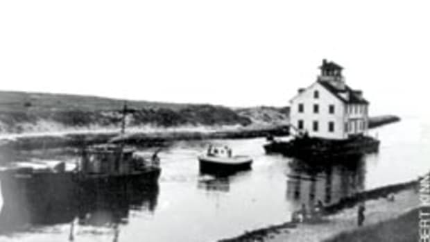 A towboat enters Menemsha Creek in summer 1952, pulling a barge carrying the new Menemsha station from Cuttyhunk, Mass.