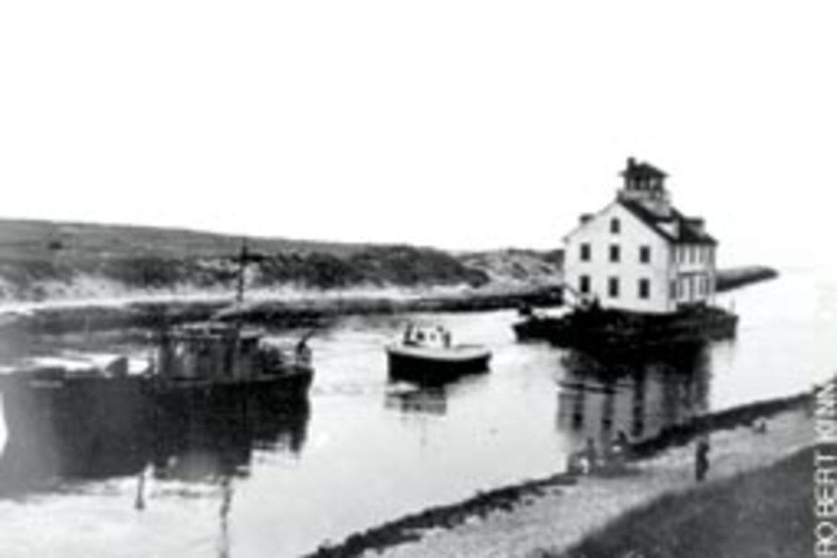 A towboat enters Menemsha Creek in summer 1952, pulling a barge carrying the new Menemsha station from Cuttyhunk, Mass.